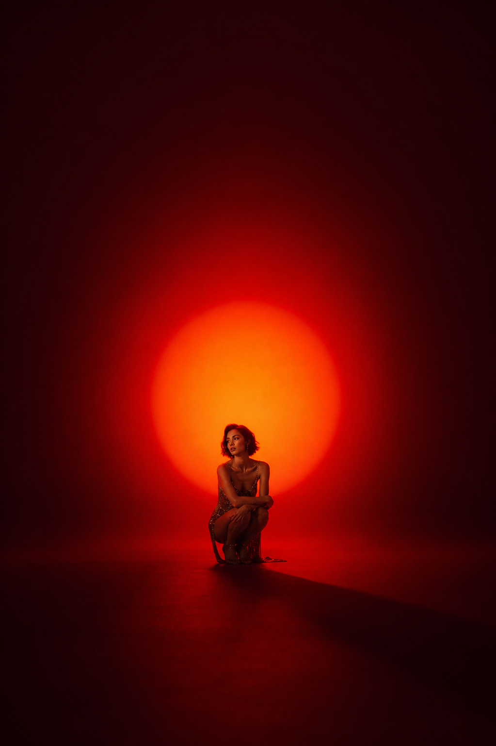 Minimalist cinematic studio portrait, ultra-high-end fashion photography, featuring a solitary woman crouched in the center of a vast seamless studio space illuminated by a dramatic glowing red-orange spotlight that radiates behind her like an artificial sunset or eclipse. The image combines contemporary editorial fashion, fine-art portraiture, and atmospheric cinematic lighting.

A young woman with a short wavy bob haircut is positioned centrally in the frame, crouching low with her arms gently crossed over her knees. Her posture feels introspective, elegant, and emotionally restrained, conveying quiet confidence and solitude. She gazes slightly off-camera into the distance, creating a contemplative and cinematic mood.

The figure occupies only a small portion of the composition, emphasizing negative space and scale. The surrounding environment feels immense, almost dreamlike, with the subject isolated inside a pool of warm light. Her silhouette is softly sculpted against the glowing backdrop, creating a powerful visual contrast between intimacy and emptiness.

She wears a delicate fashion-forward outfit with subtle metallic embellishments that catch the light. The wardrobe remains understated, allowing the lighting and composition to dominate the image. Soft highlights define the contours of her shoulders, arms, and face.

Behind her, a massive circular red-orange light source creates a radiant halo effect that gradually fades into deep crimson and near-black shadows at the edges of the frame. The seamless cyclorama studio environment disappears into darkness, creating a surreal void-like atmosphere. A long soft-edged shadow extends across the floor, adding depth and cinematic tension.

Composition:

Vertical portrait orientation
Extreme negative space
Subject positioned near the lower center of the frame
Large glowing circular light centered behind the model
Minimalist fine-art composition
Small human figure contrasted against vast empty space
Symmetrical balance with subtle asymmetrical body pose
Editorial magazine aesthetic

Lighting:

Single giant spotlight projection
Deep red, crimson, and orange color wash
Artificial sunset effect
Soft diffusion around light source
Dramatic falloff into darkness
Subtle rim lighting around hair and shoulders
Cinematic shadow casting across floor
High contrast yet soft transitions
Atmospheric haze enhancing light bloom

Color Palette:

Deep crimson red
Burnt orange
Amber glow
Scarlet highlights
Dark burgundy shadows
Warm skin tones
Black and near-black gradients
Monochromatic warm spectrum

Mood & Atmosphere:

Dreamlike
Solitary
Introspective
Cinematic
Melancholic elegance
Contemporary fine art
Fashion editorial minimalism
Quiet emotional tension
Surreal stage-like environment
Modern gallery installation aesthetic

Photography Style References:
James Turrell-inspired light installation, Nadav Kander atmosphere, Tim Walker minimalism, contemporary Vogue editorial, fashion campaign photography, cinematic portraiture, luxury beauty campaign, fine-art studio photography, modern visual poetry.

Technical Specifications:
Medium-format digital camera, Hasselblad X2D, 80mm lens, ultra-shallow depth of field, studio cyclorama, volumetric light diffusion, soft atmospheric haze, HDR tonal range, photorealistic skin texture, cinematic color grading, subtle film grain, luxury editorial quality, museum-grade fine-art photography, 8K resolution.

Keywords:
minimalist fashion portrait, red spotlight, glowing halo backdrop, cinematic studio lighting, dramatic negative space, luxury editorial photography, atmospheric portrait, fine-art fashion image, contemporary visual art, surreal lighting installation, emotional portraiture, modern magazine aesthetic.

Negative Prompt:
crowded scene, multiple people, busy background, props, furniture, harsh facial shadows, overexposed highlights, low-resolution, cartoon style, CGI look, excessive makeup, distracting elements, text, logos, watermarks, cluttered composition, bright daylight, outdoor environment.